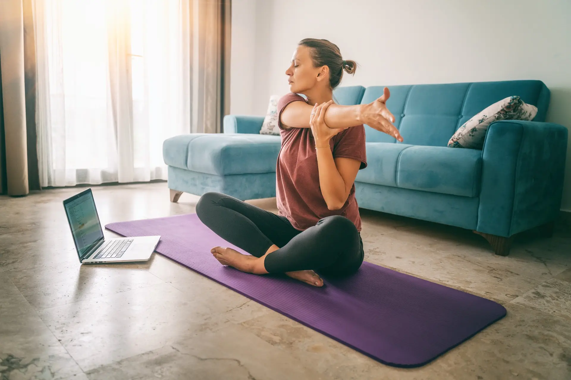 Woman practicing a seated shoulder stretch at home beside a laptop